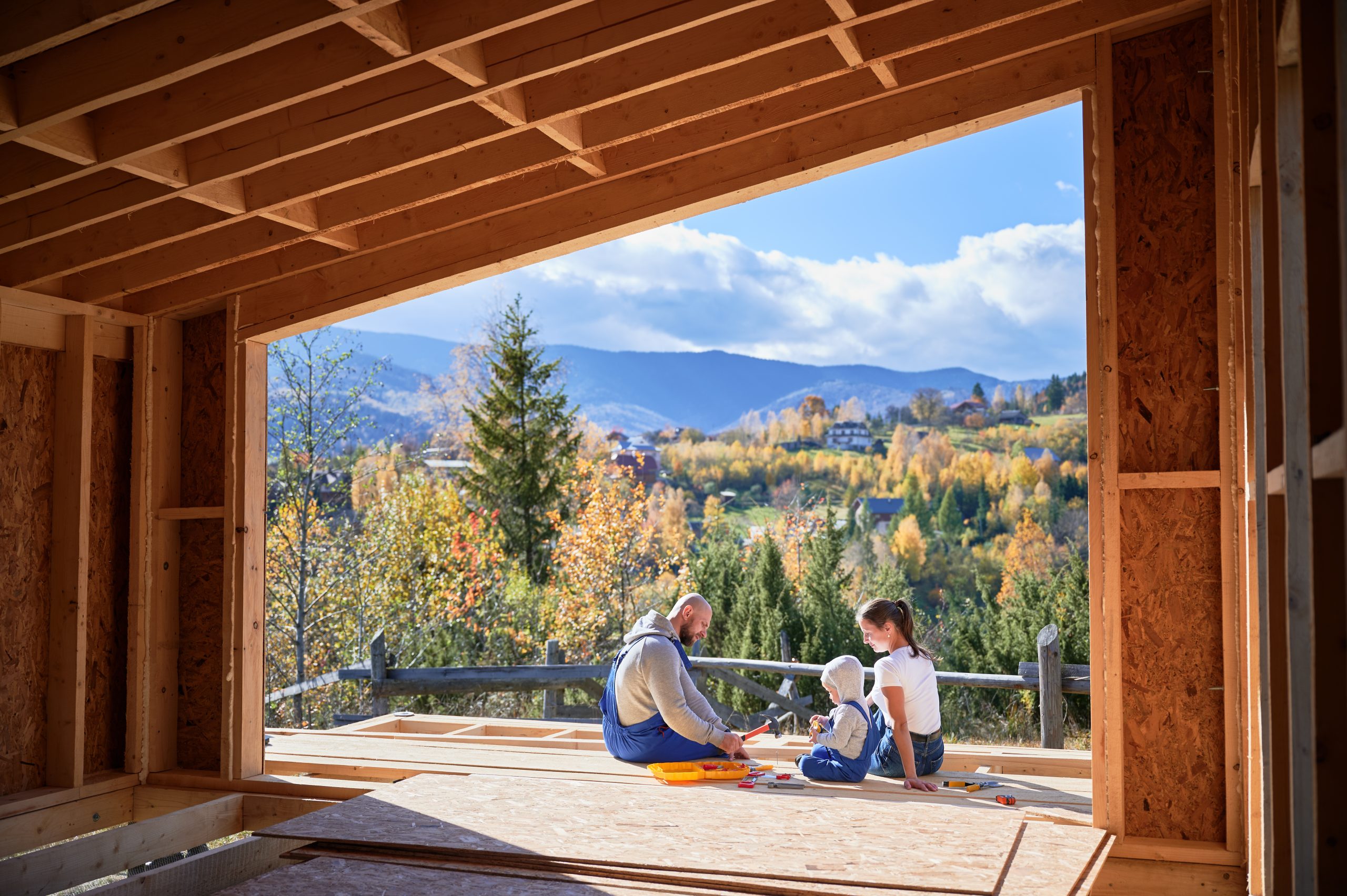Father, mother and son building wooden frame house. Toddler boy helping his daddy, while woman looking for them on construction site. View from inside a house. Carpentry, family concept.
